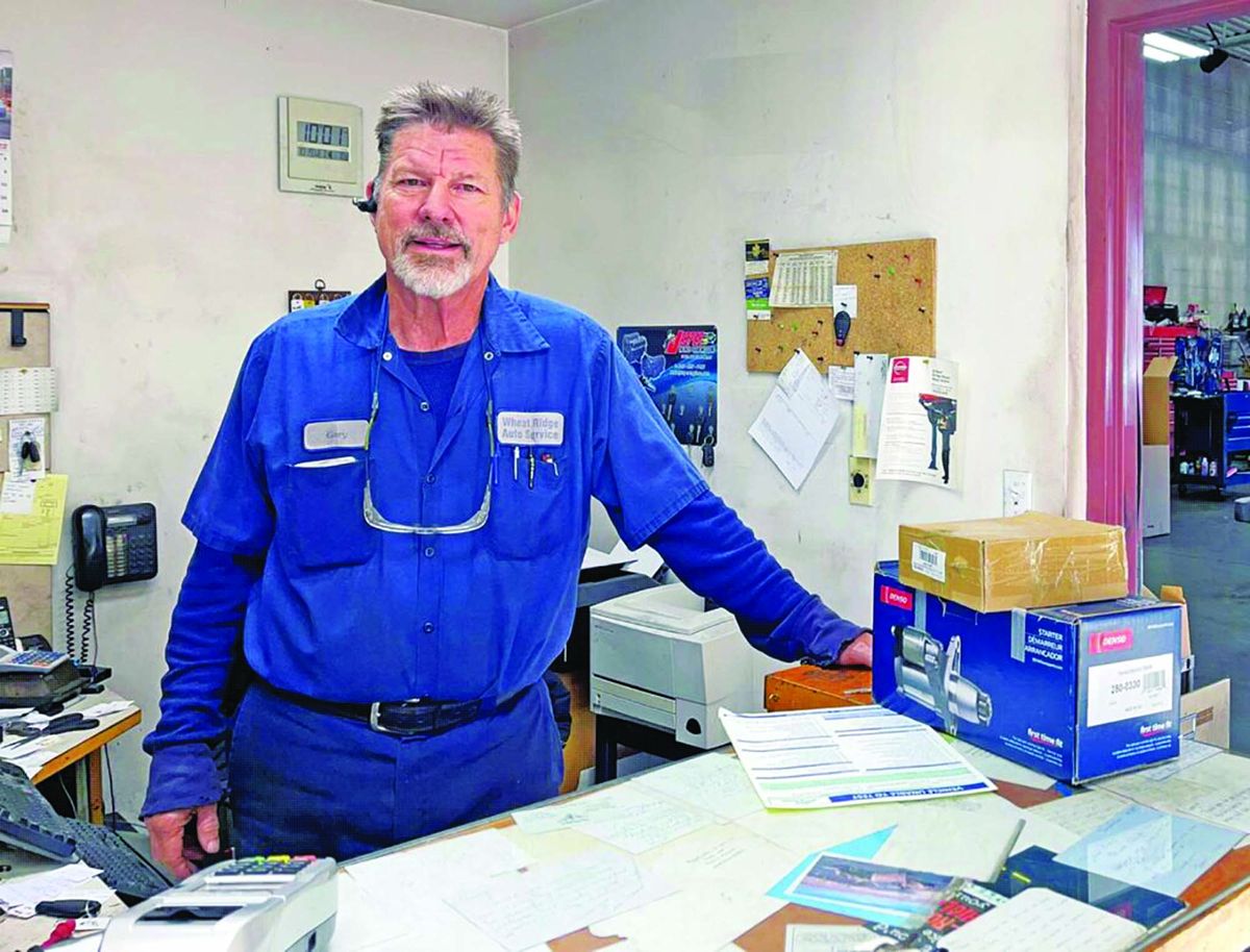 Gary Bryan, in a blue Wheat Ridge Auto Service work shirt with his name tag, standing at the shop’s front counter
